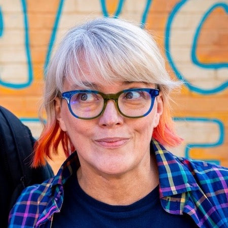 Portrait of the author with blond and sunset coloured hair wearing glasses with blue-green frames. T-shirt says 'I came to make Conservatives uncomfortable'.